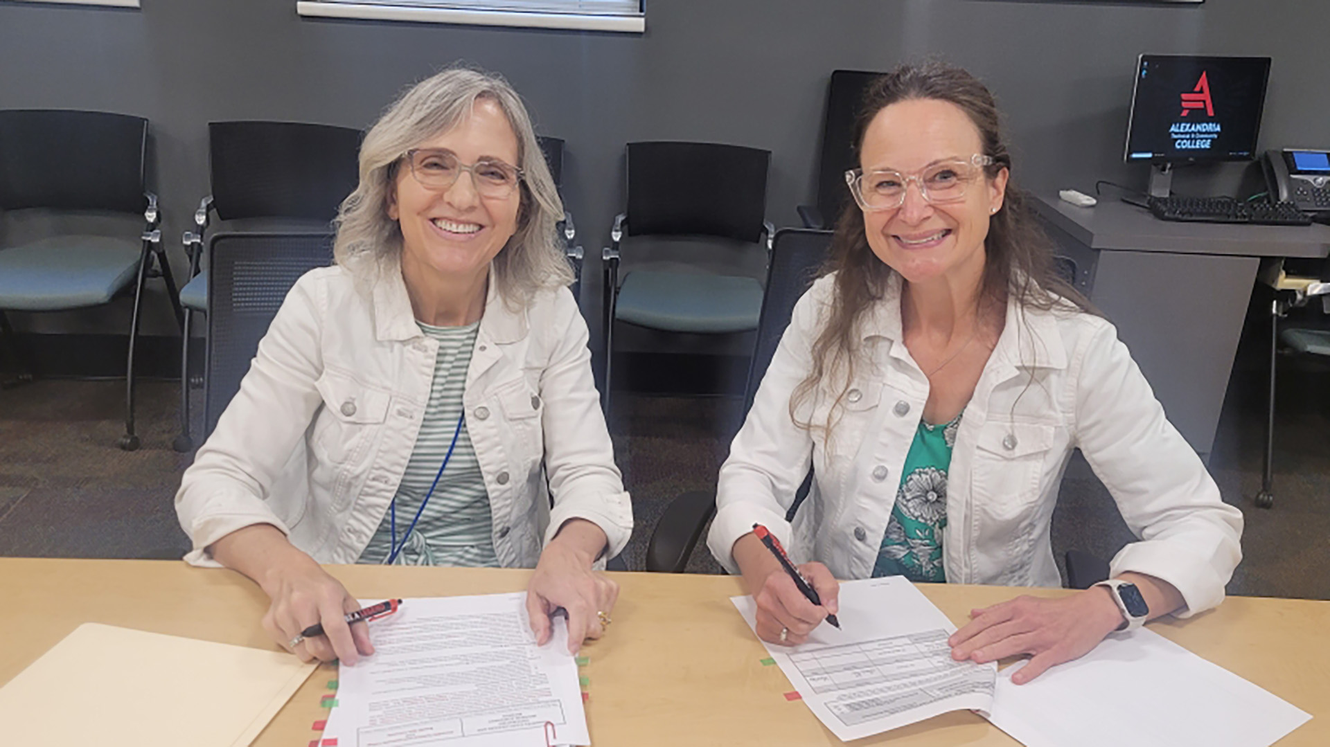 Two women wearing identical white denim jackets are signing paperwork on a table.