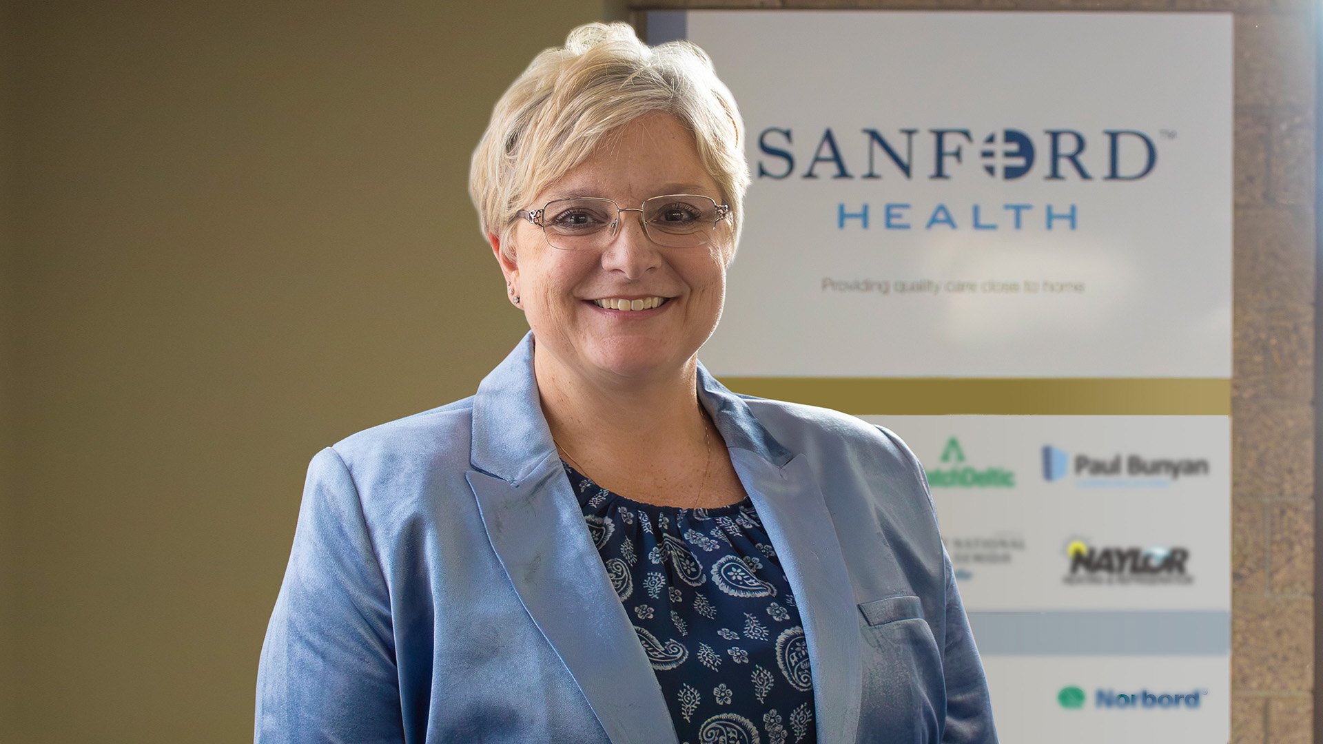 A blonde haired woman with glasses is wearing a shiny, light blue blazer over a dark blue shirt. A Sanford Health logo is visible on the wall behind her.