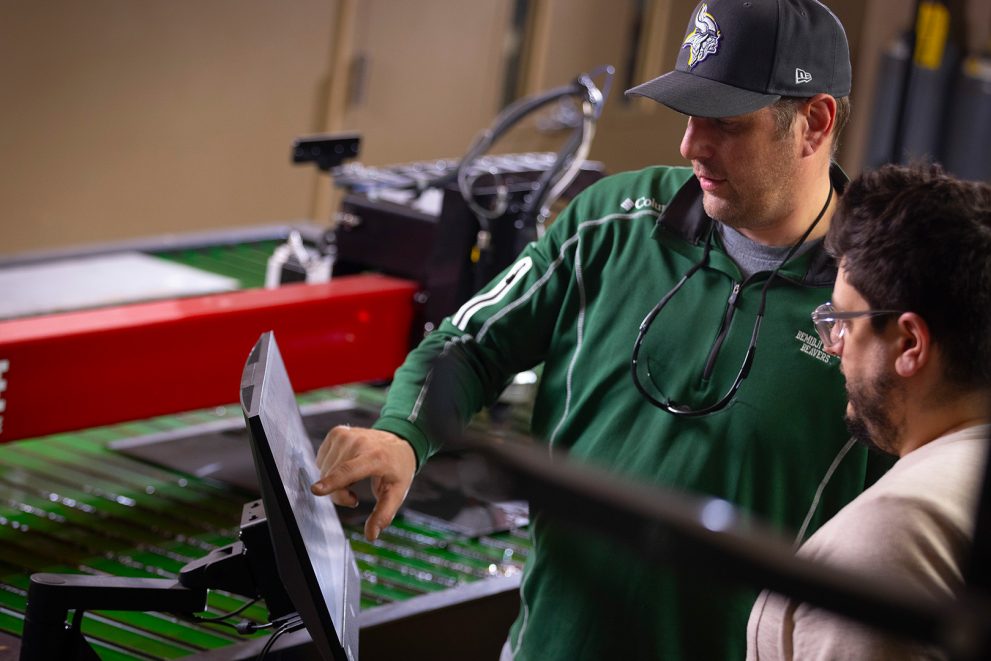 Two people work on a touchscreen positioned in front of a table-based plasma cutter. A man in a green nylon pullover and a green hat with the BSU athletics logo is touching the screen while a bearded man wearing glasses and a beige shirt watches on from the side.