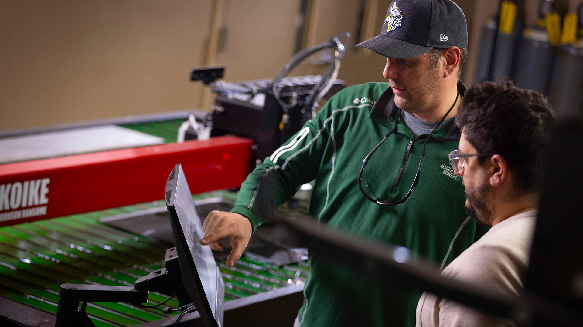 Two people work on a touchscreen positioned in front of a table-based plasma cutter. A man in a green nylon pullover and a green hat with the BSU athletics logo is touching the screen while a bearded man wearing glasses and a beige shirt watches on from the side.