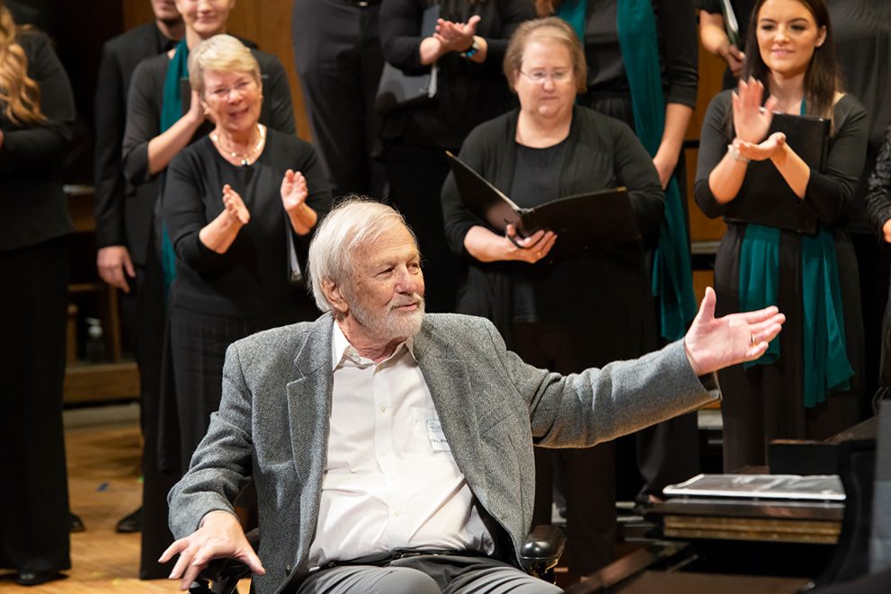 A photo of Dr. Paul Brandvik at a concert celebrating BSU choir's 85th anniversary in 2022. He has white hair and wears a grey suit coat with a white shirt underneath. He is seated, and waving to a crowd wiht his right hand. Behind him, a group of BSU choir alumni are applauding him.