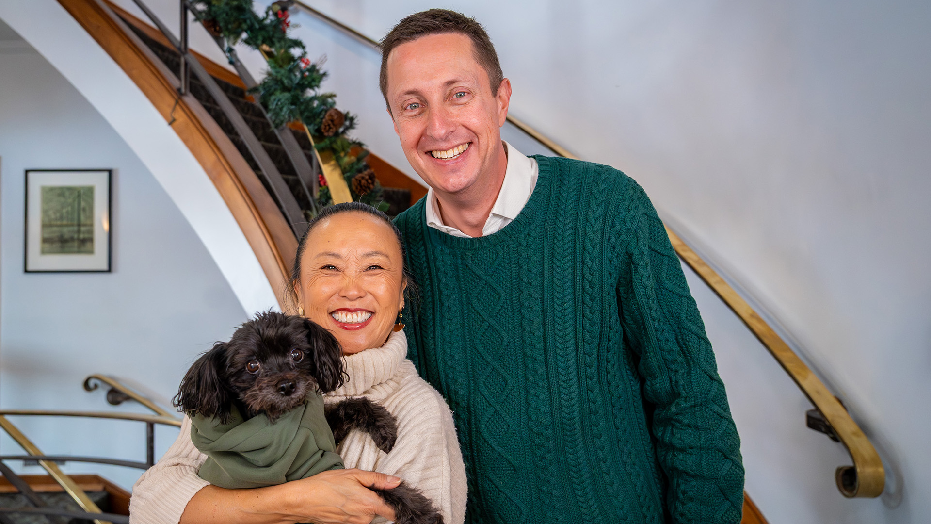BSU President Dr. John L. Hoffman., wearing a green sweater, and his spouse Dr. Joy Hoffman, wearing a beige sweater, are by the stairs in BSU's David Park House. Joy is holding their small black dog, Jackson.