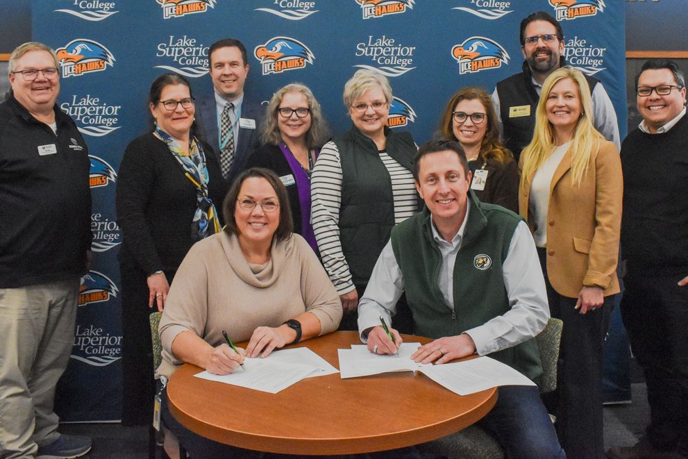 LSC President and BSU President John L. Hoffman are sitting together at a round table signing documents. Standing behind them are a number of other representatives from both campuses.