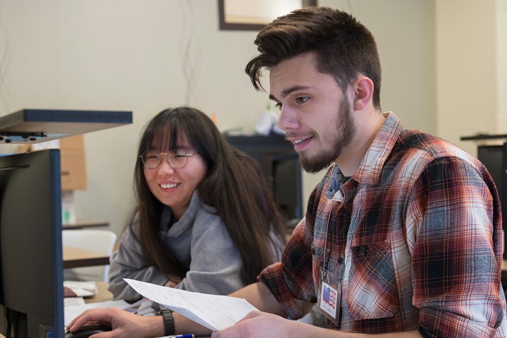 A BSU student in a red, black and white plaid shirt works at a computer with a woman in glasses wearing a grey sweatshirt.
