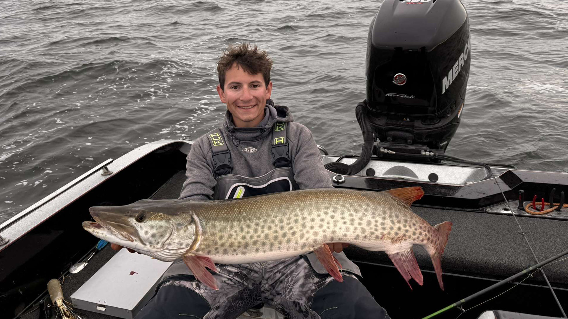 BSU grad student Baylor Short sits at the back of a boat with a large Mercury outboard motor behind him. He is smiling and holding a large muskie in front of him with both hands.
