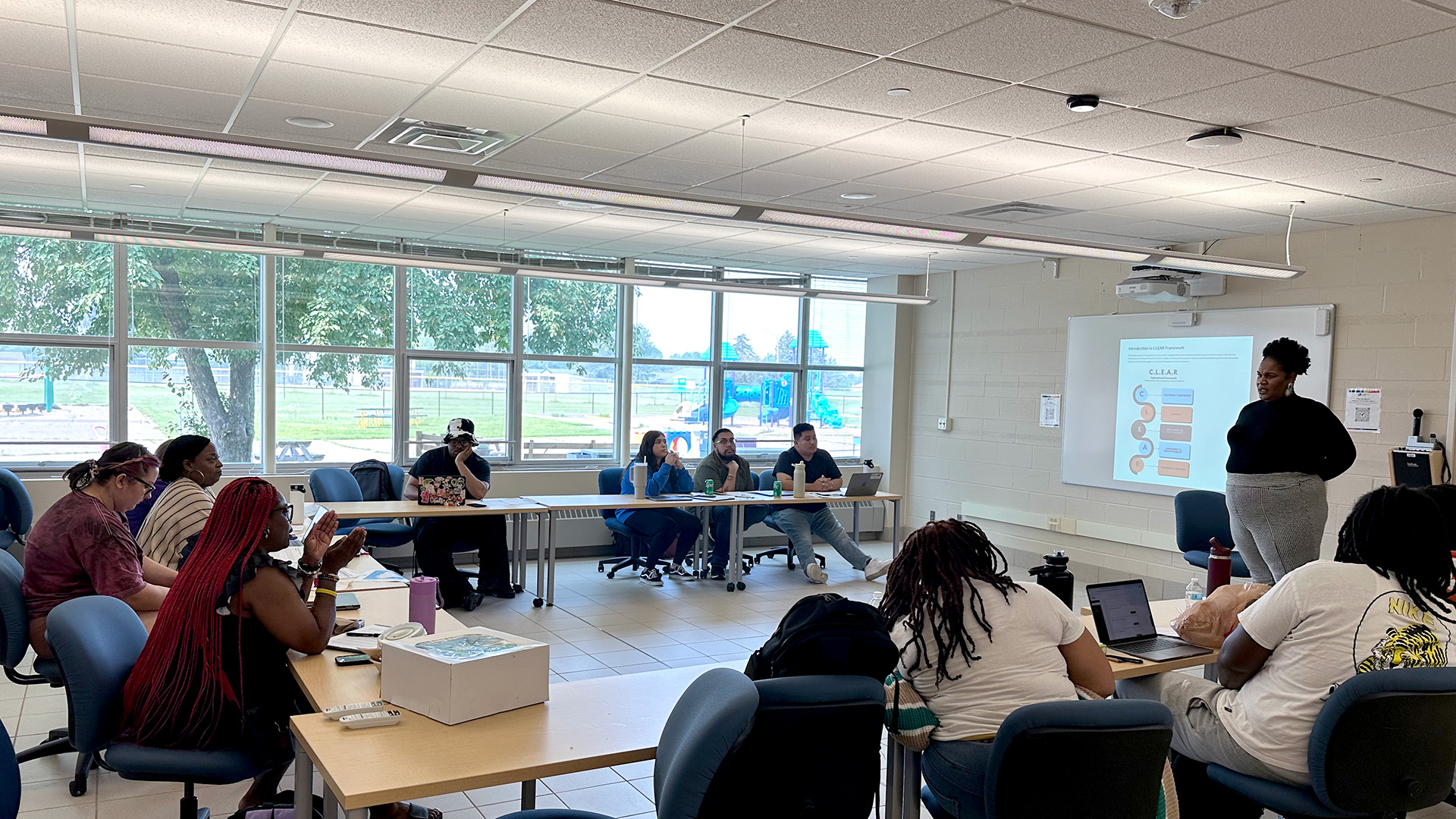 A woman gives a presentation to a group of students seated at tables. A PowerPoint presentation is on a screen behind her.