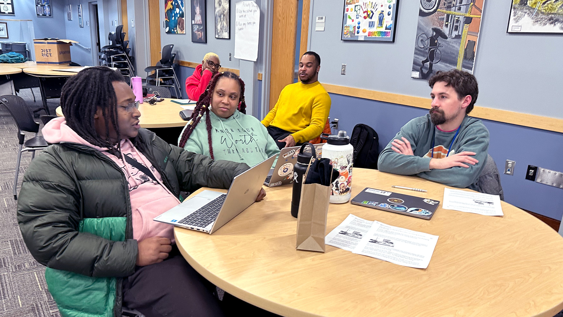 Three people are sitting at a round table having a conversation; two of them have laptops open. A fourth person at a table behind them is also participating.