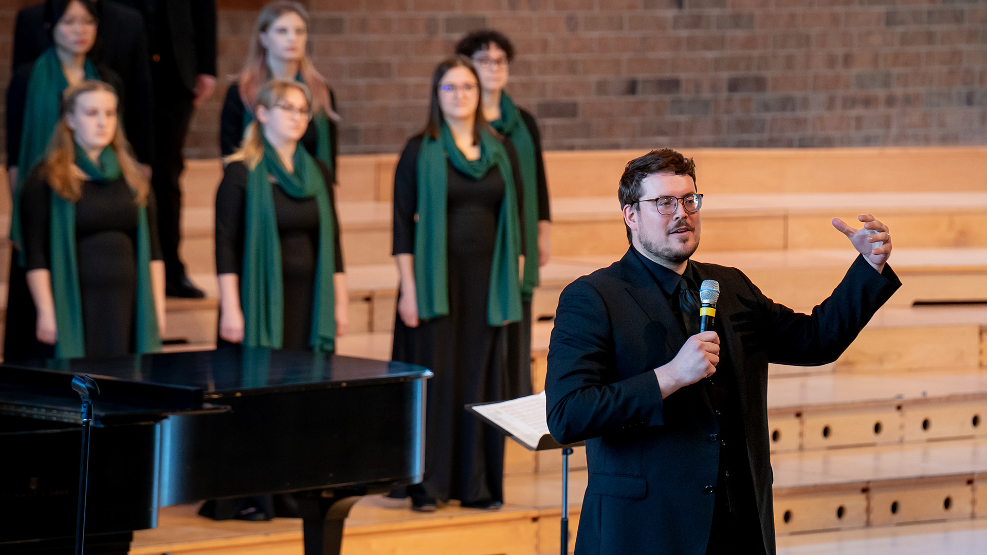 Dr. Isaac Lovdahl holds a microphone and speaks to a crowd, with the Bemidji Choir behind him, at the 2025 ACDA Minnesota Conference.