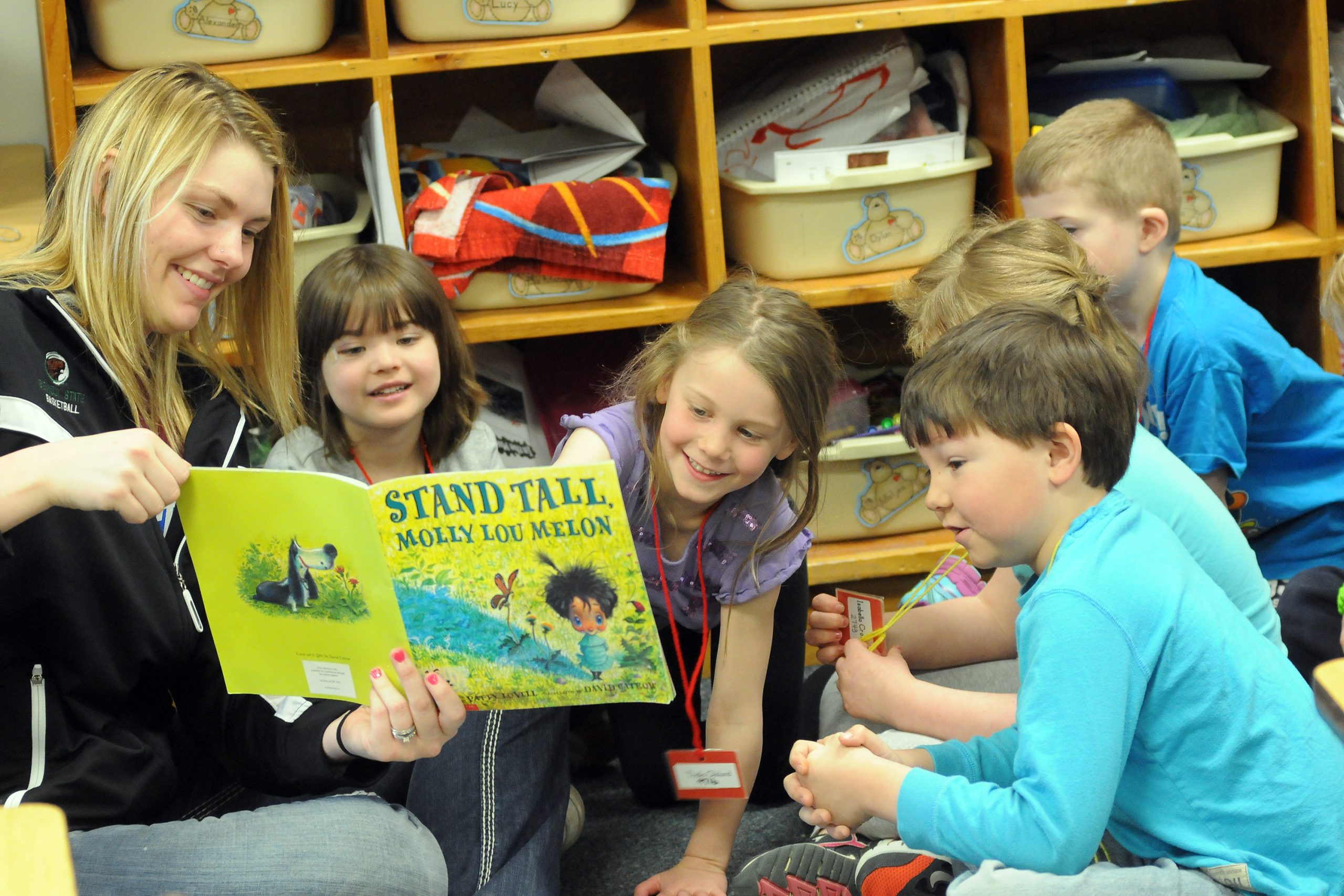 Teacher reading to a group of students from a book called "Stand Tall" by Molly Lou Melon