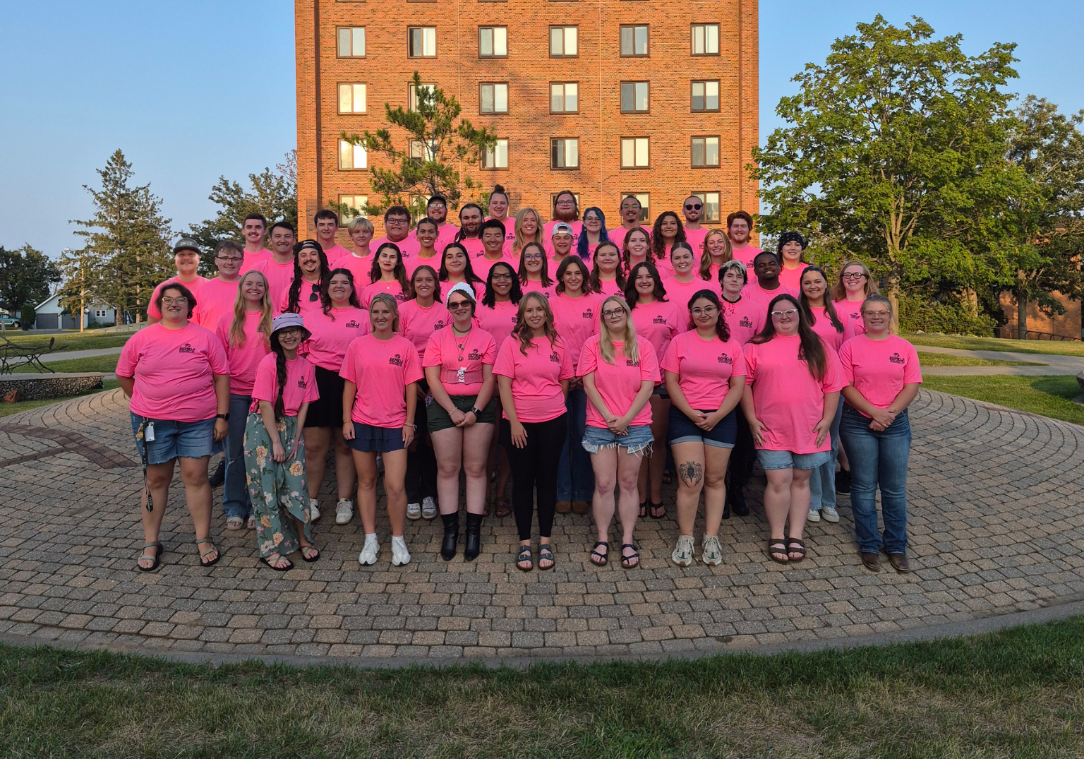 Large group wearing matching bright pink shirts poses in front of a tall red-brick building.
