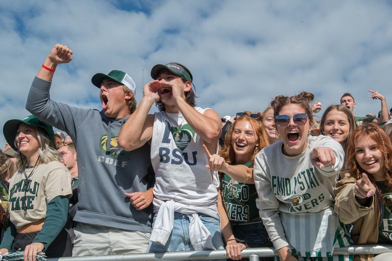 Students wearing Bemidji State gear are cheering outside a football game against a backdrop of a cloudy but blue sky