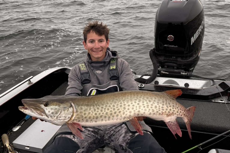 BSU grad student Baylor Short sits at the back of a boat with a large Mercury outboard motor behind him. He is smiling and holding a large muskie in front of him with both hands.