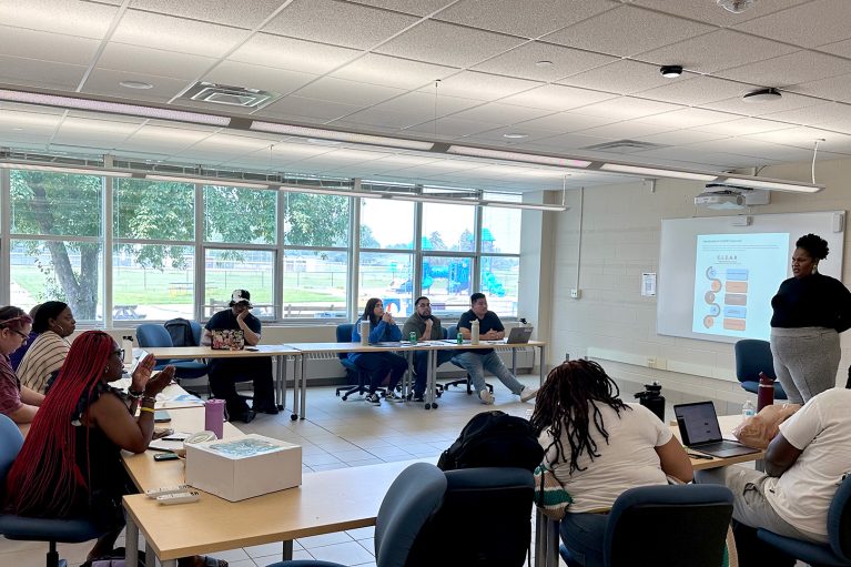A woman gives a presentation to a group of students seated at tables. A PowerPoint presentation is on a screen behind her.