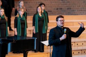 Dr. Isaac Lovdahl holds a microphone and speaks to a crowd, with the Bemidji Choir behind him, at the 2025 ACDA Minnesota Conference.